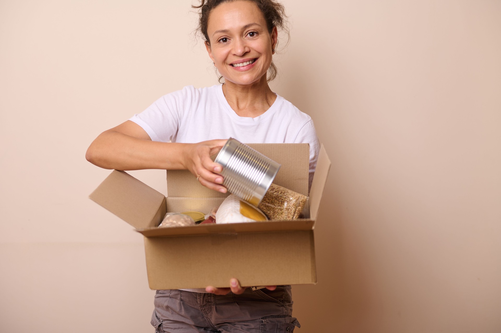 Smiling volunteer woman wearing white t-shirt, packing goods into a mockup cardboard box, preparing humanitarian help for charity and donation. Food Bank Day.